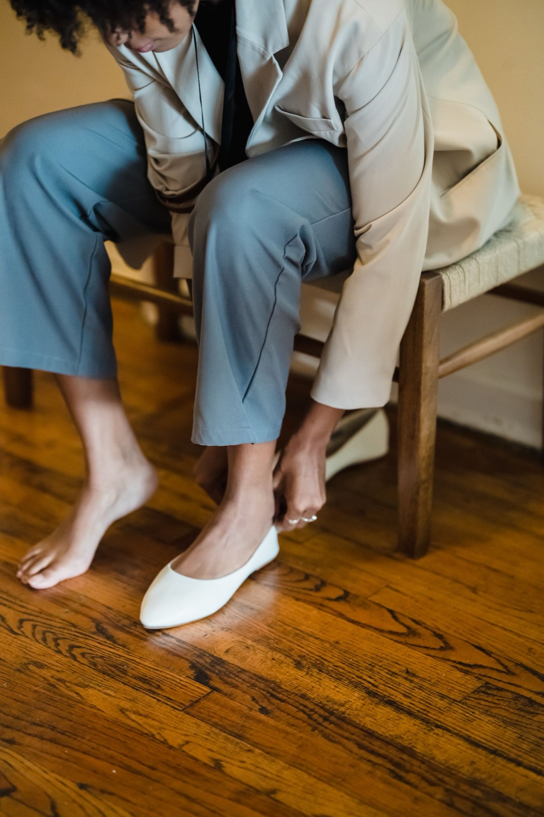 A woman in a beige blazer and gray trousers sitting on a woven bench, fastening a white ballet flat on one foot