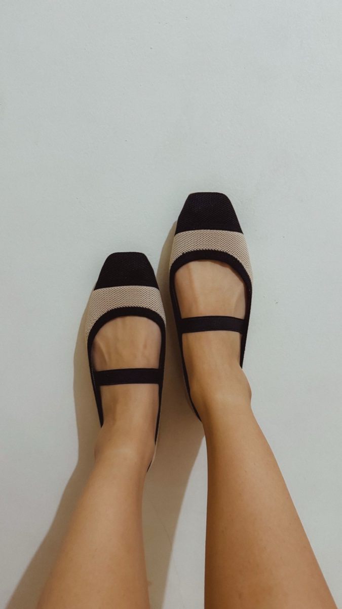 Feet in two-tone knit ballet flats with black elastic mary jane straps and black square toes, photographed from above against a white wall