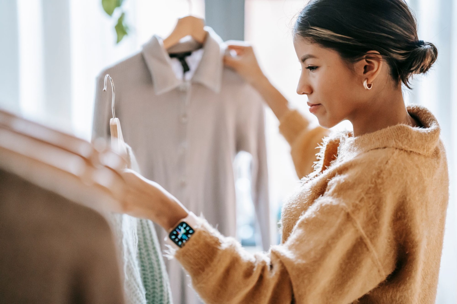 Woman in a tan sweater holding up two hangers and comparing garments near a bright window in a clothing store