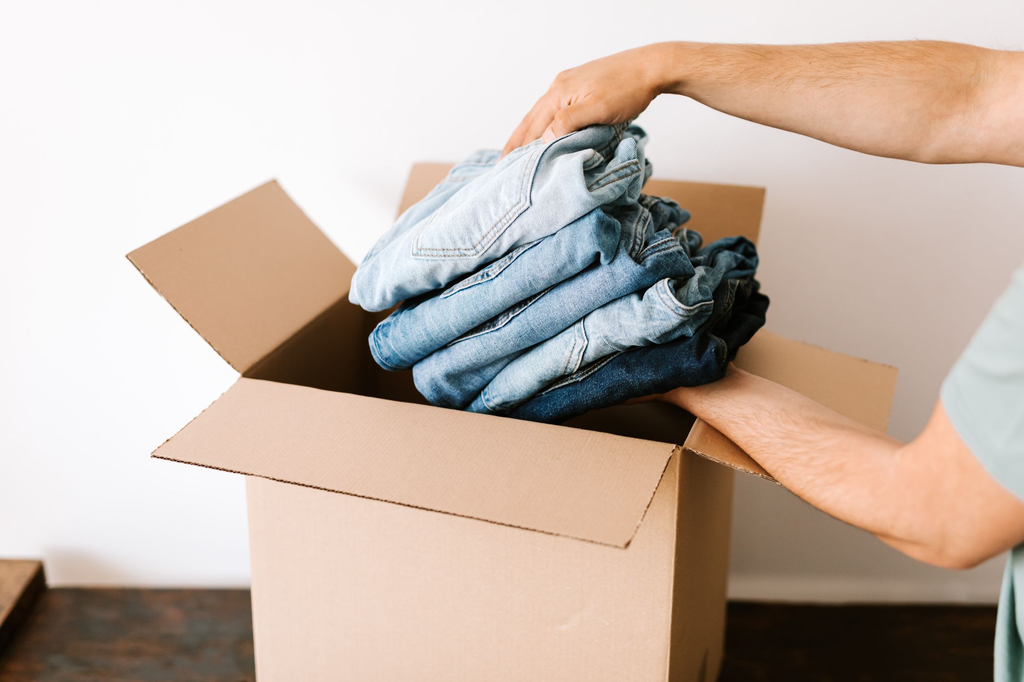 A person folding a stack of jeans into an open cardboard box against a white wall