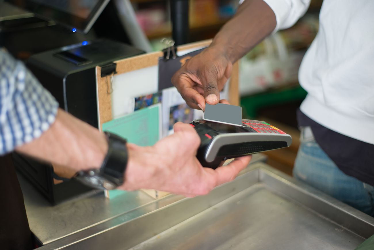 A shopper's hand extending a gray card toward a payment terminal held by a cashier at a store counter