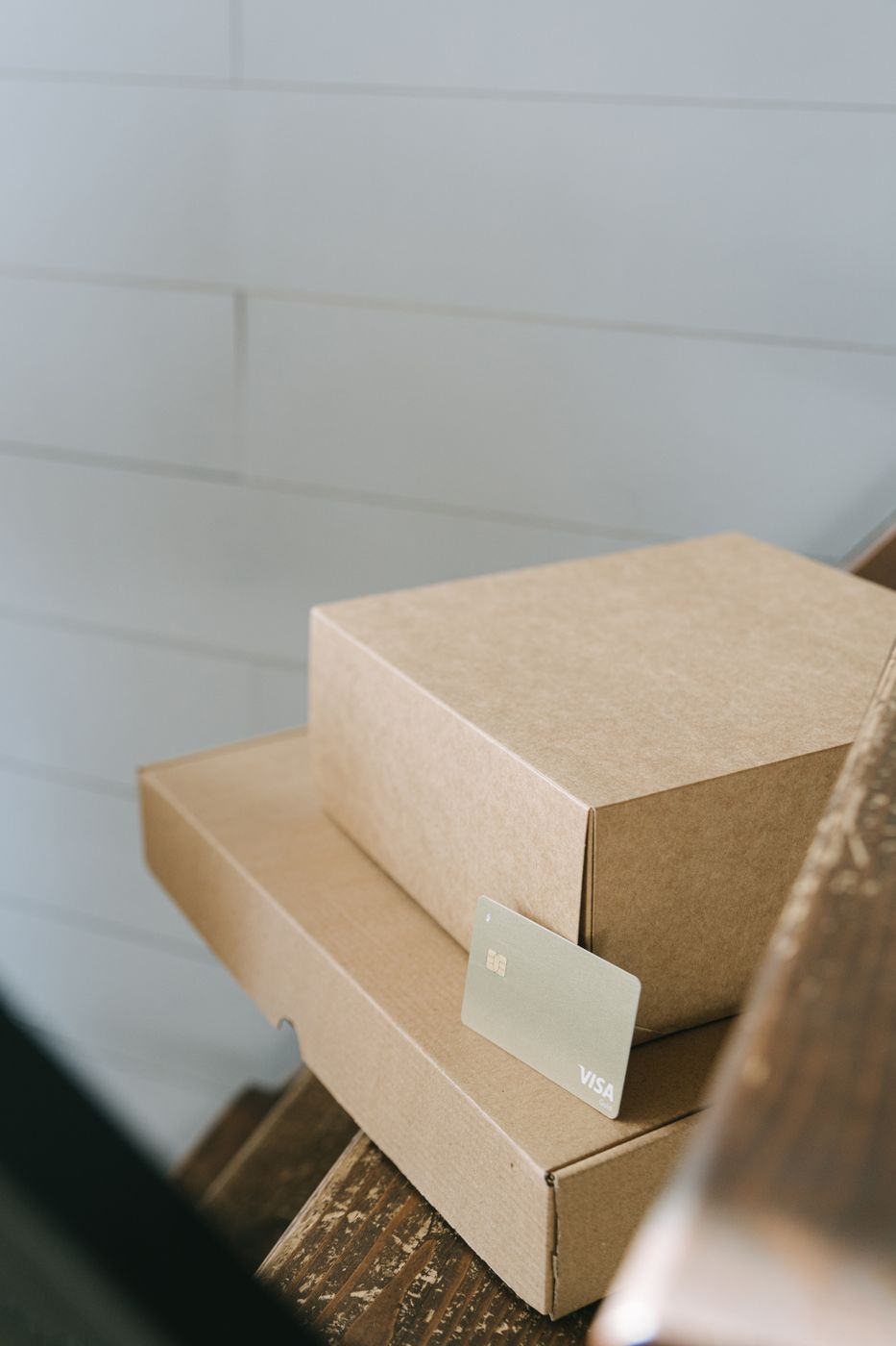 A payment card resting against two stacked plain cardboard mailer boxes on wooden stairs beside a white wall