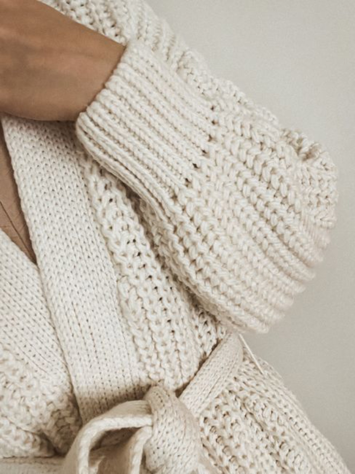 Close-up of a cream chunky hand-knit cardigan sleeve with thick ribbed cuff, a hand resting on the shoulder