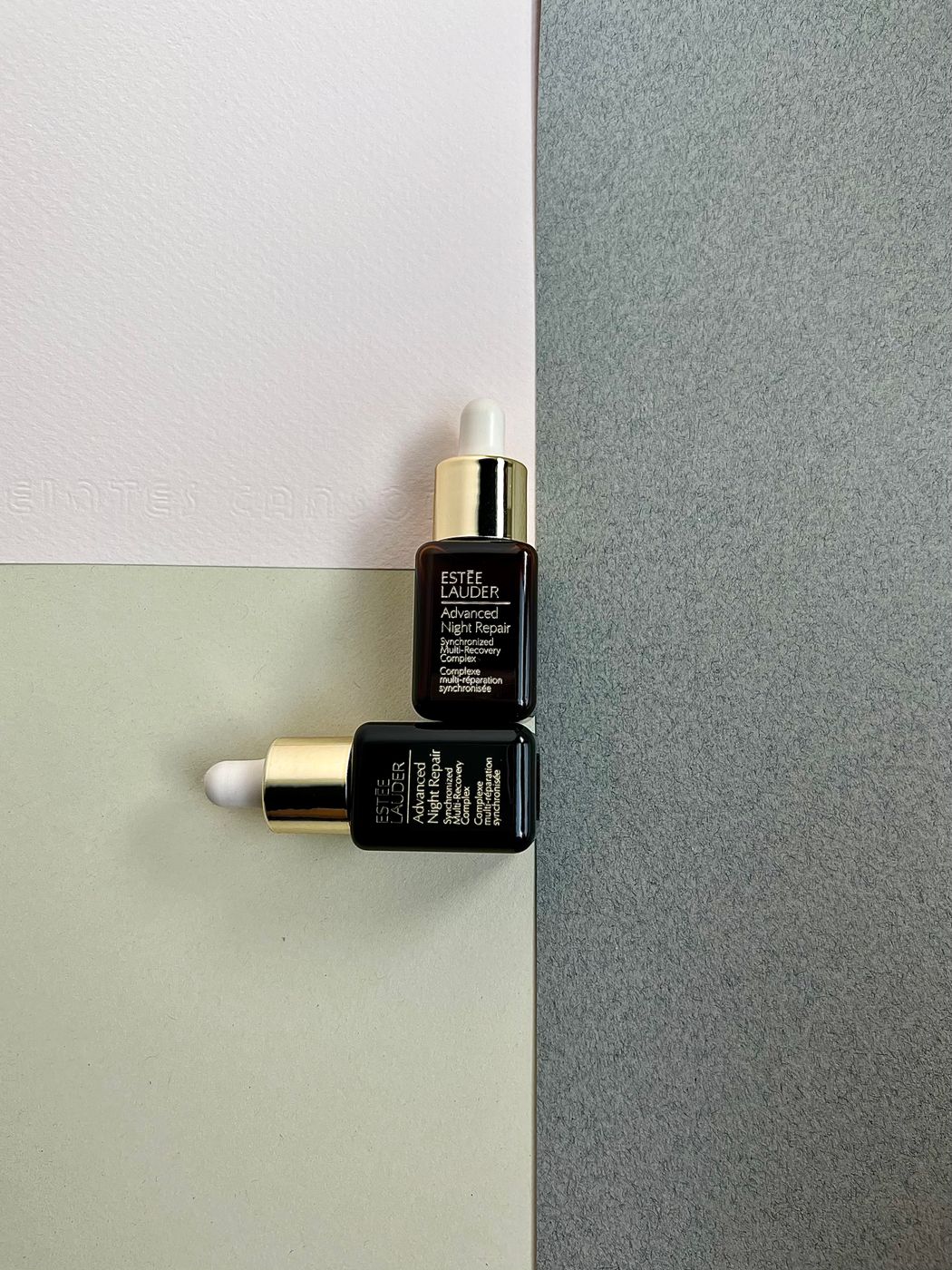 Two small dark Estee Lauder Advanced Night Repair dropper bottles with gold collars resting on a pale two-tone background