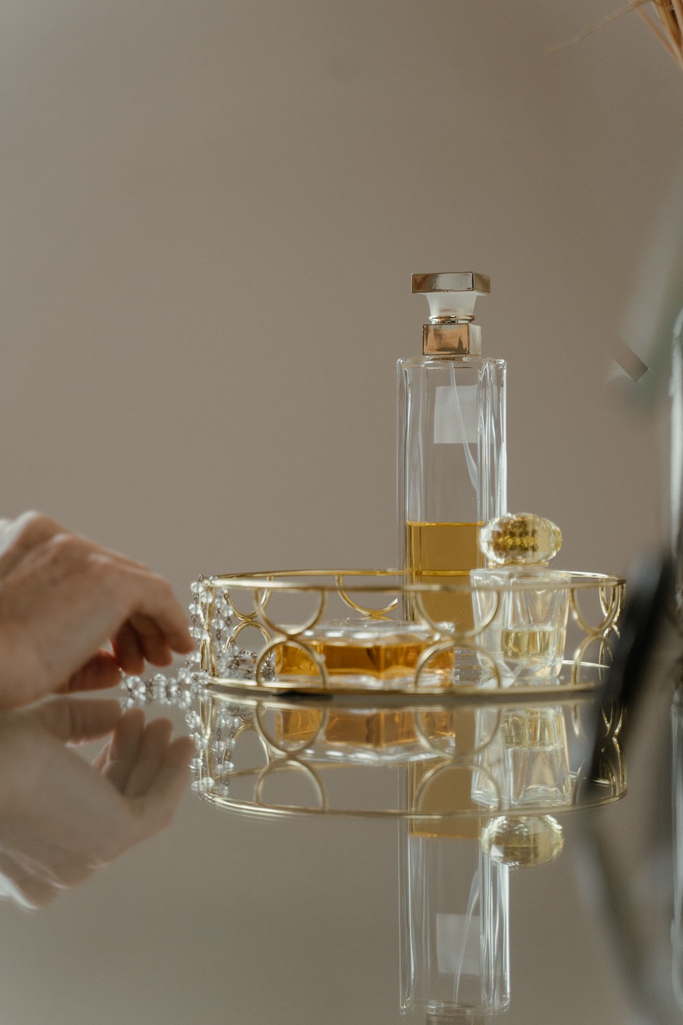 A gold wire tray on a mirrored table holding three filled glass perfume bottles, with a hand reaching toward a beaded chain at the left