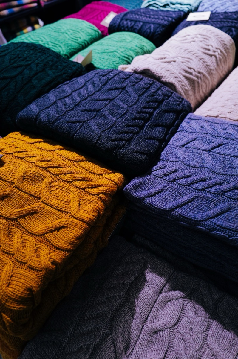 Cable-knit sweaters folded in tall stacks on a display table, mustard, navy, lilac and green, with paper swing tags still attached