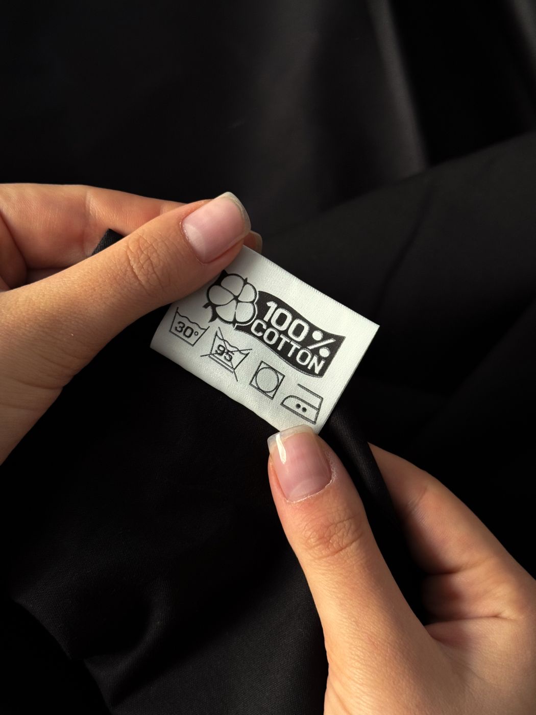 Two hands holding a small white care label printed 100% cotton with wash symbols, against dark fabric