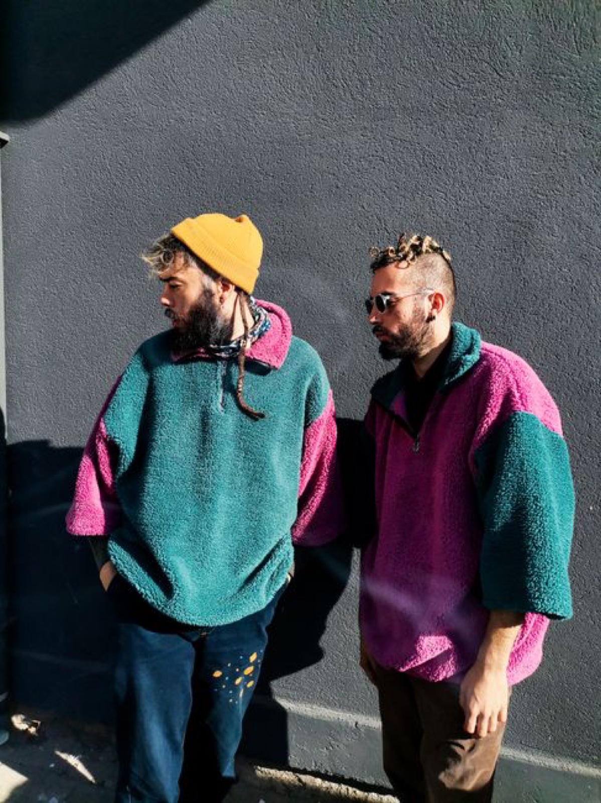 Two men in oversized teal and magenta colorblock deep-pile fleece pullovers standing against a dark gray wall in hard sunlight