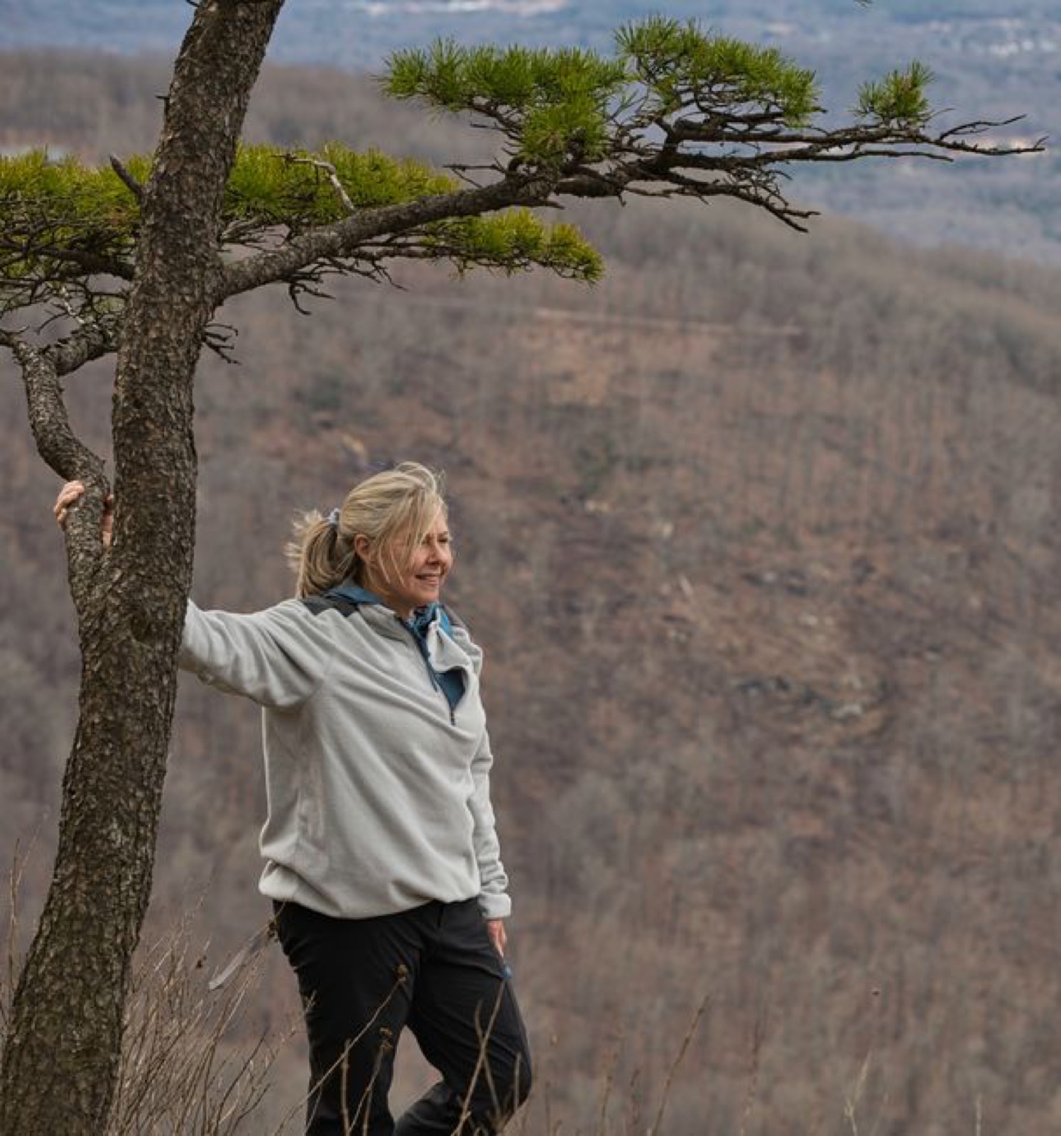A woman in a light gray fleece pullover standing by a pine tree above a valley on an overcast hike