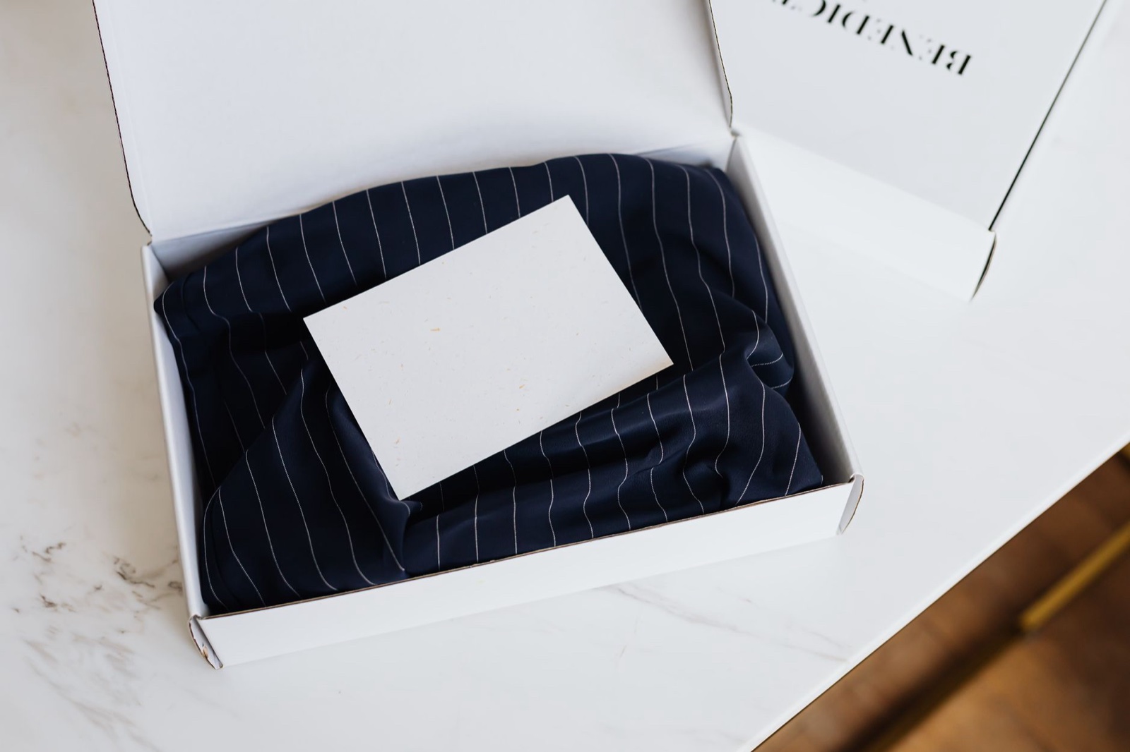 An open white cardboard box holding a folded navy pinstripe garment with a blank cream card resting on top
