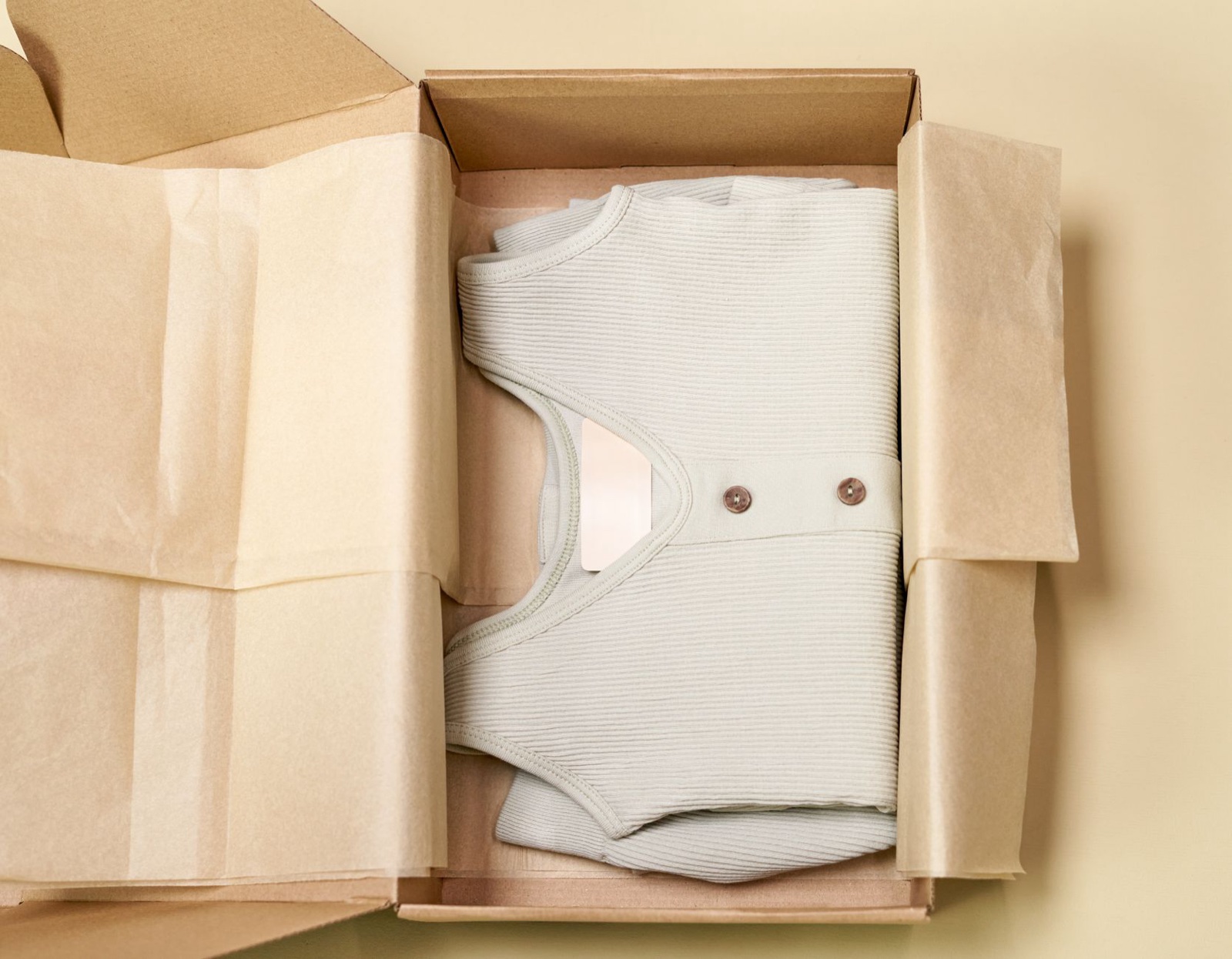 Overhead view of a ribbed cream knit top folded inside an open cardboard mailer lined with kraft tissue paper