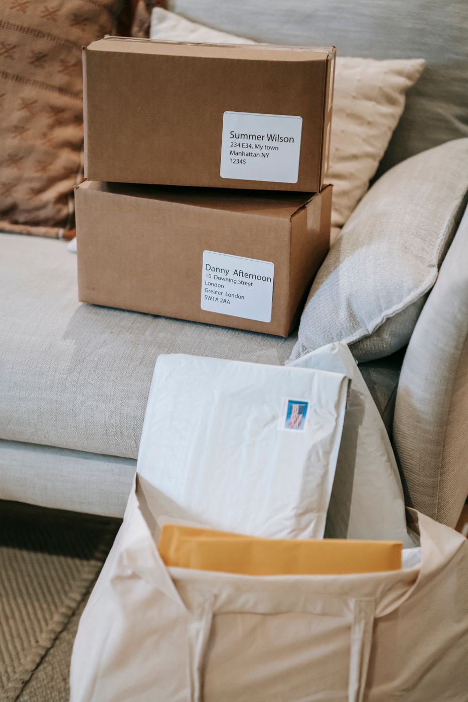 Two cardboard parcels with printed address labels stacked on a sofa beside a fabric tote holding mailing envelopes