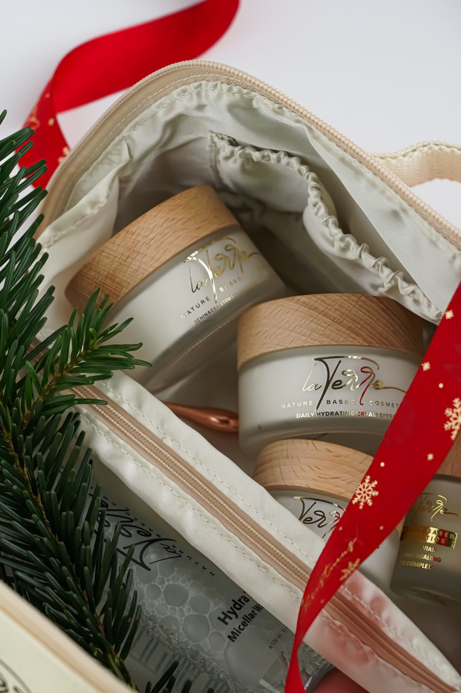 Festive skincare gift pouch holding three branded cream jars, with red ribbon and a pine sprig alongside
