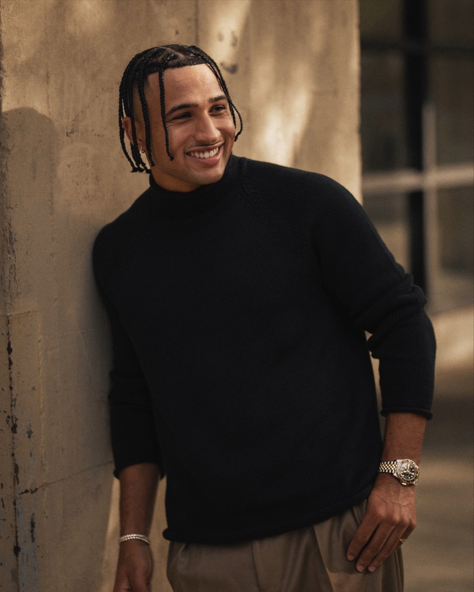 A man in a black classic rollneck sweater and beige trousers leaning against a stone wall in warm evening light