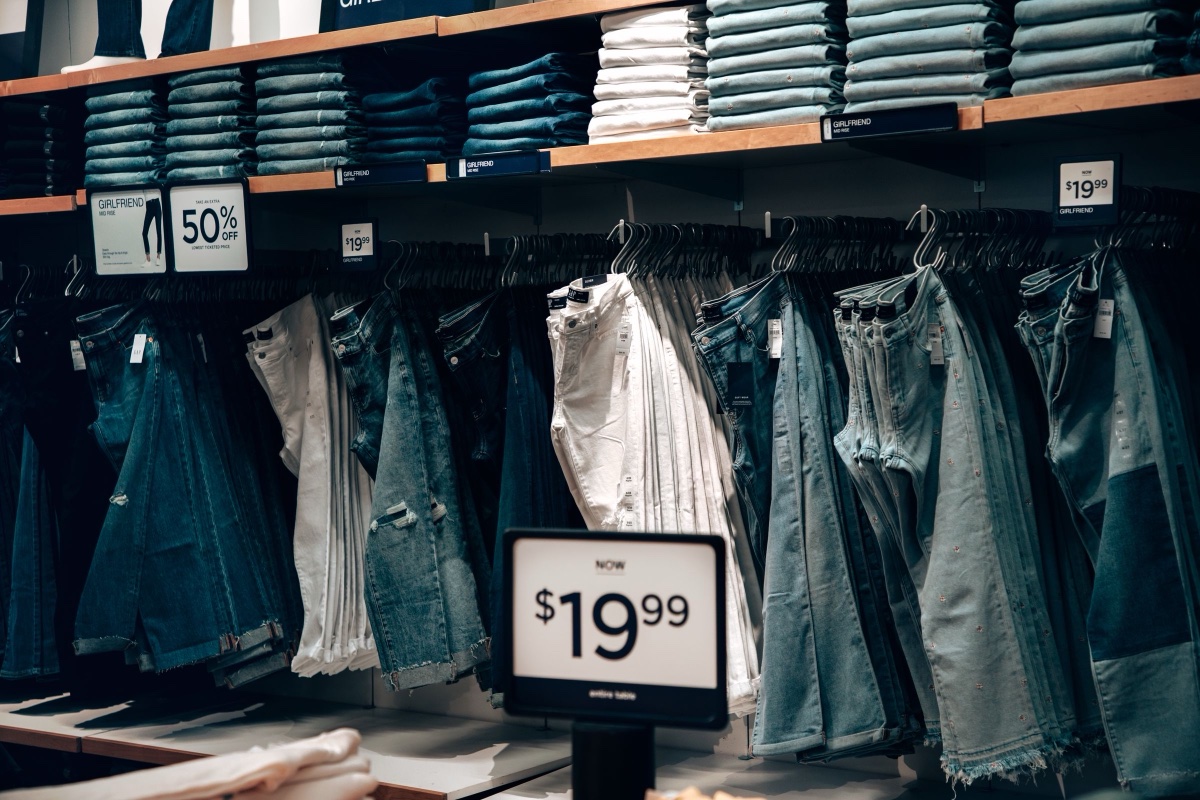 A store's denim wall, folded jeans on a top shelf and hanging pairs below, with a 50 percent off card and a now $19.99 sign clipped to the rail