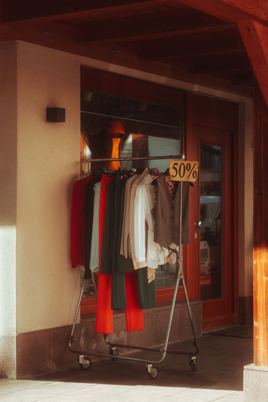 An outdoor clothing rail parked beside a shop door in warm evening light, with a hand-lettered cardboard sign reading 50% clipped to the rail