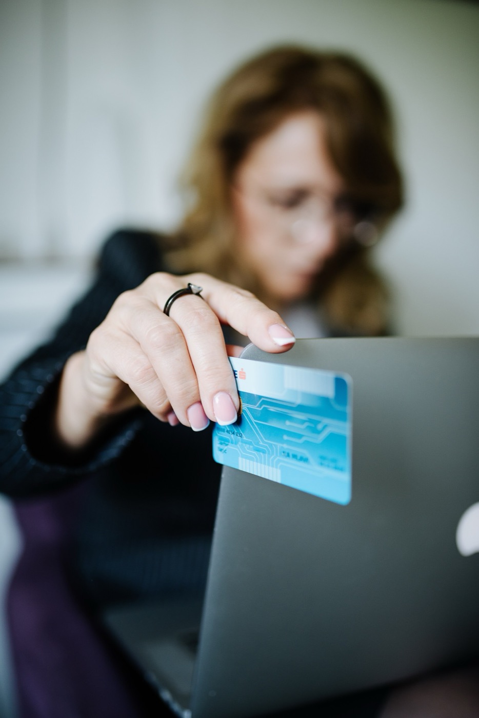 A person at a laptop holds a blue payment card up over the screen edge, face out of focus behind it