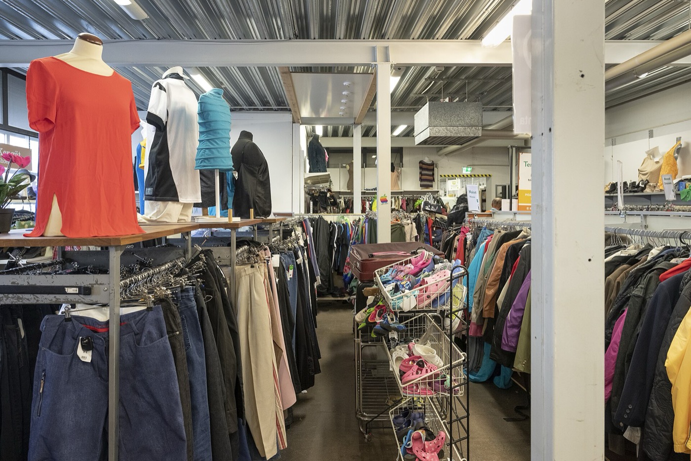 A second-hand clothing shop floor with packed rails of jackets and trousers, mannequins on a table and wire trolleys of shoes down the centre aisle