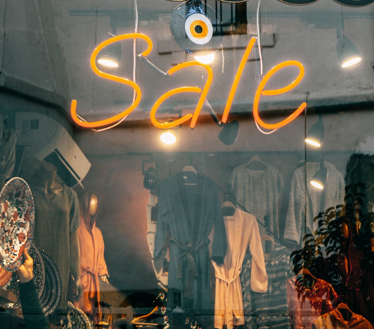 An orange neon sign spelling the word Sale in cursive hanging inside a shop window with clothing racks visible behind the glass