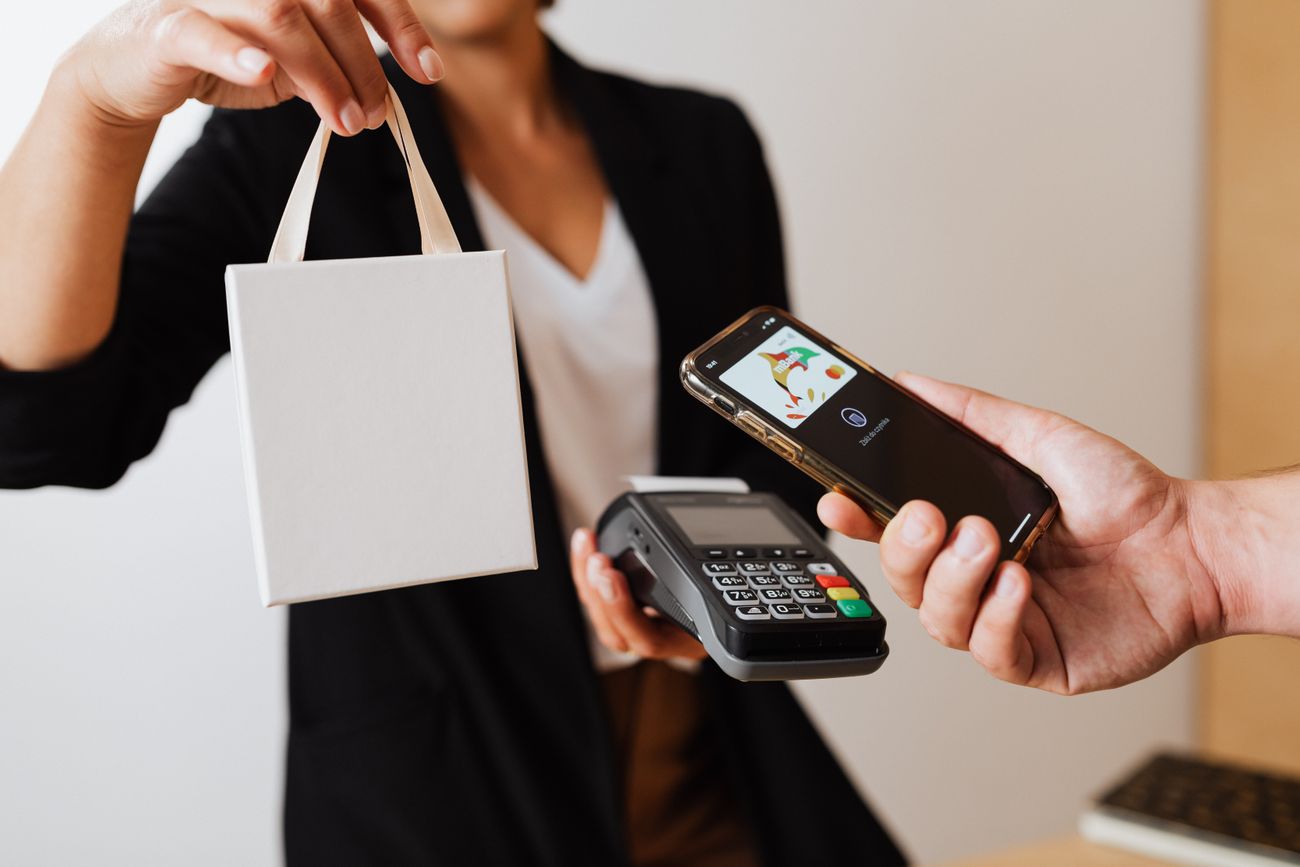 A store clerk handing over a small white shopping bag while a customer pays by phone at a terminal