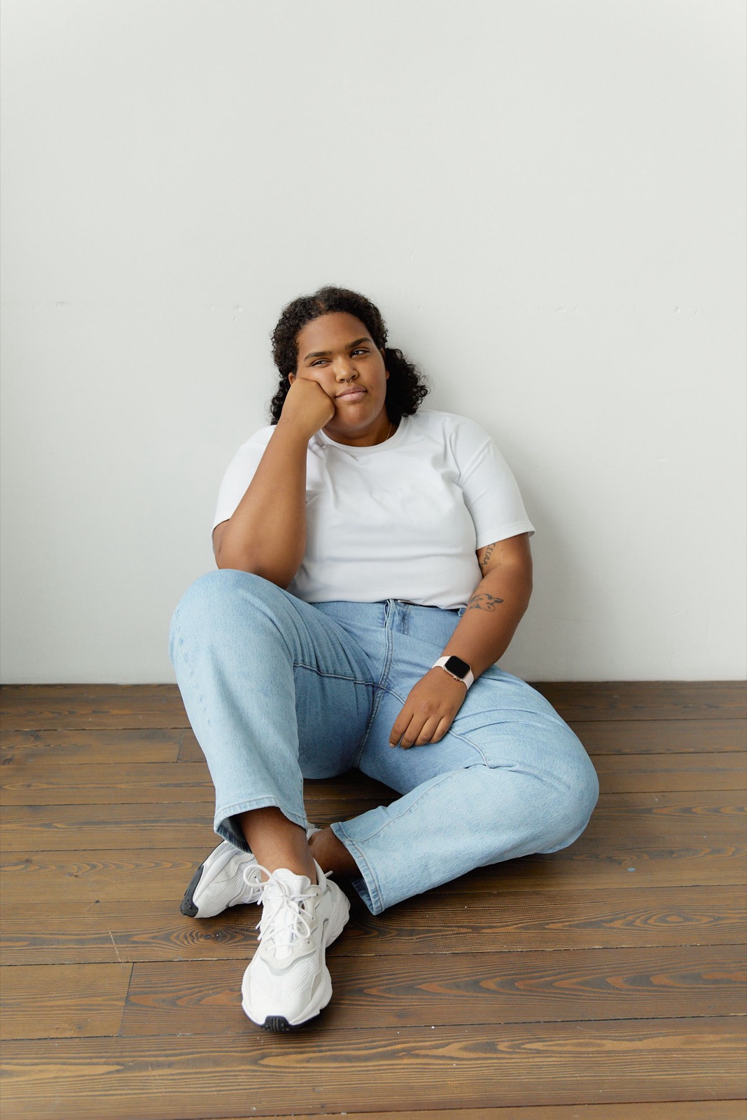 Plus-size woman in a white t-shirt and light wash jeans sitting on a wooden floor against a white wall