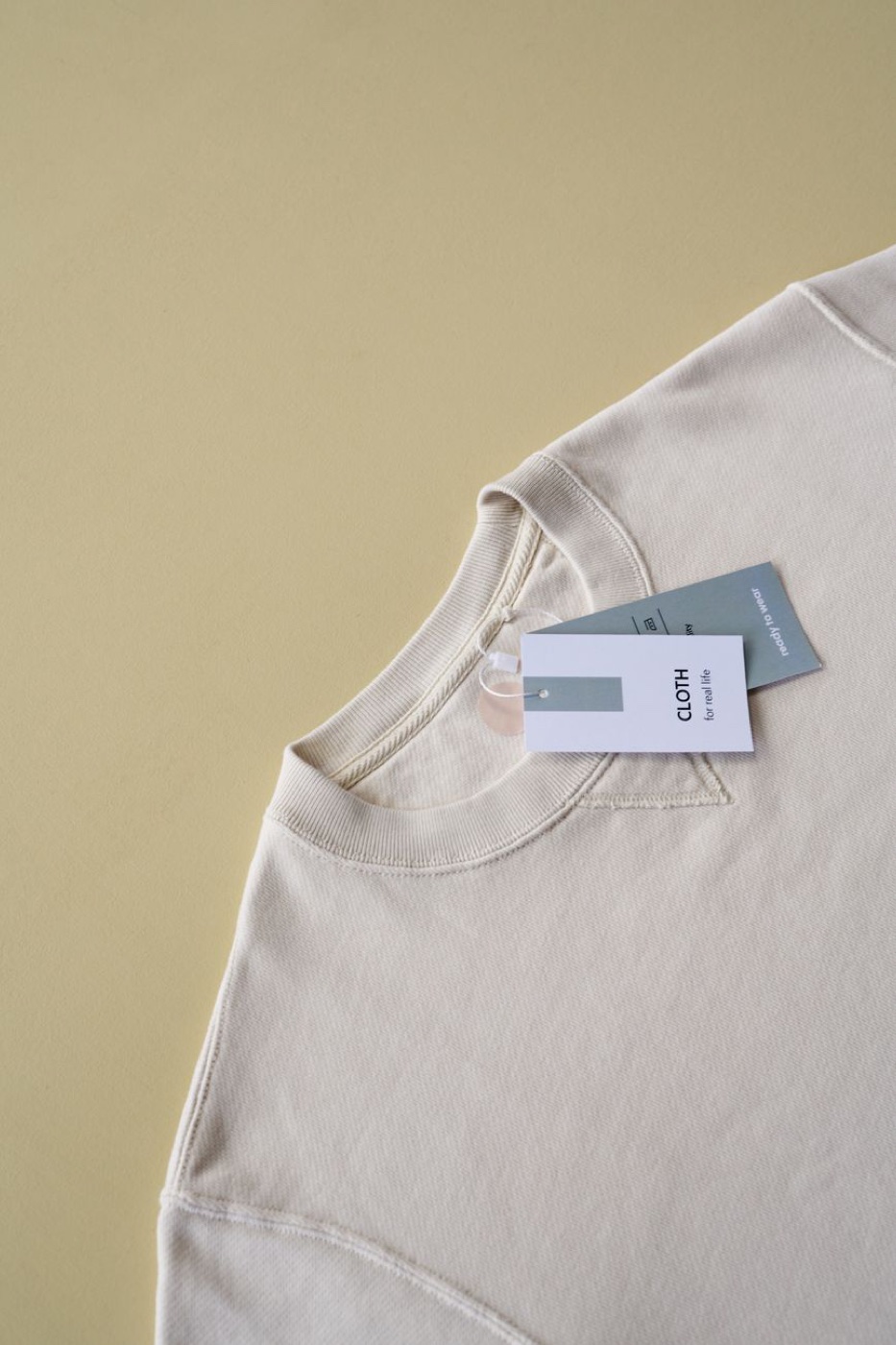 Two paper swing tags reading CLOTH for real life and ready to wear, attached to the neckline of a cream sweatshirt on a pale yellow surface