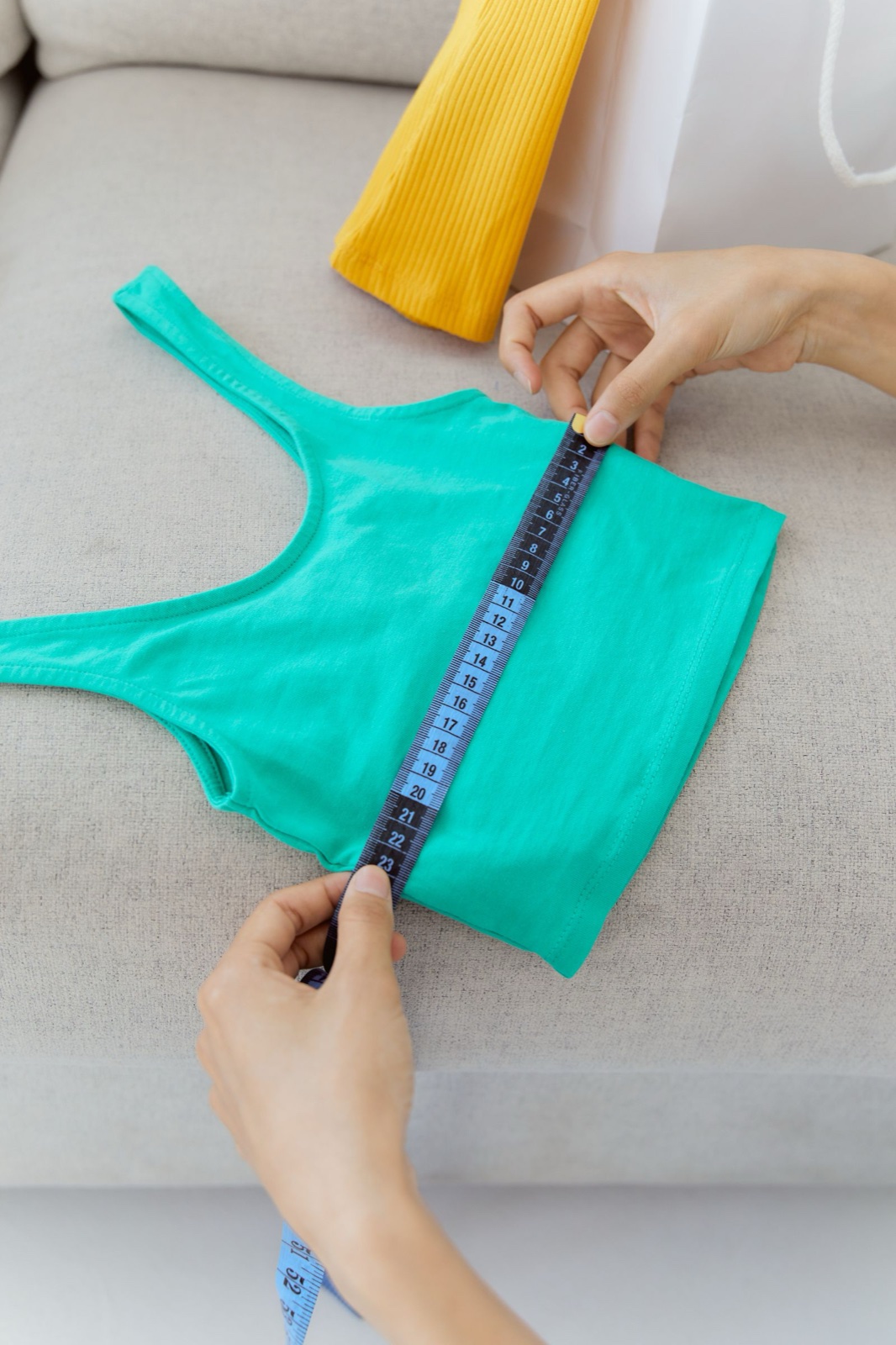 Hands running a tape measure across a teal tank top laid flat on a gray sofa cushion