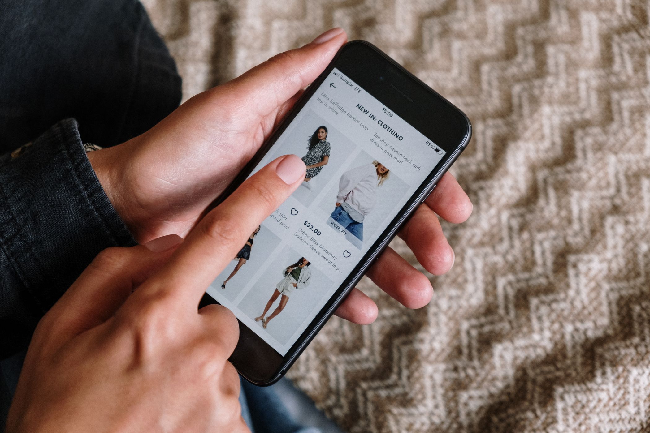 Two hands holding a smartphone showing a clothing app listing page with dresses, a price of thirty-two dollars and heart icons