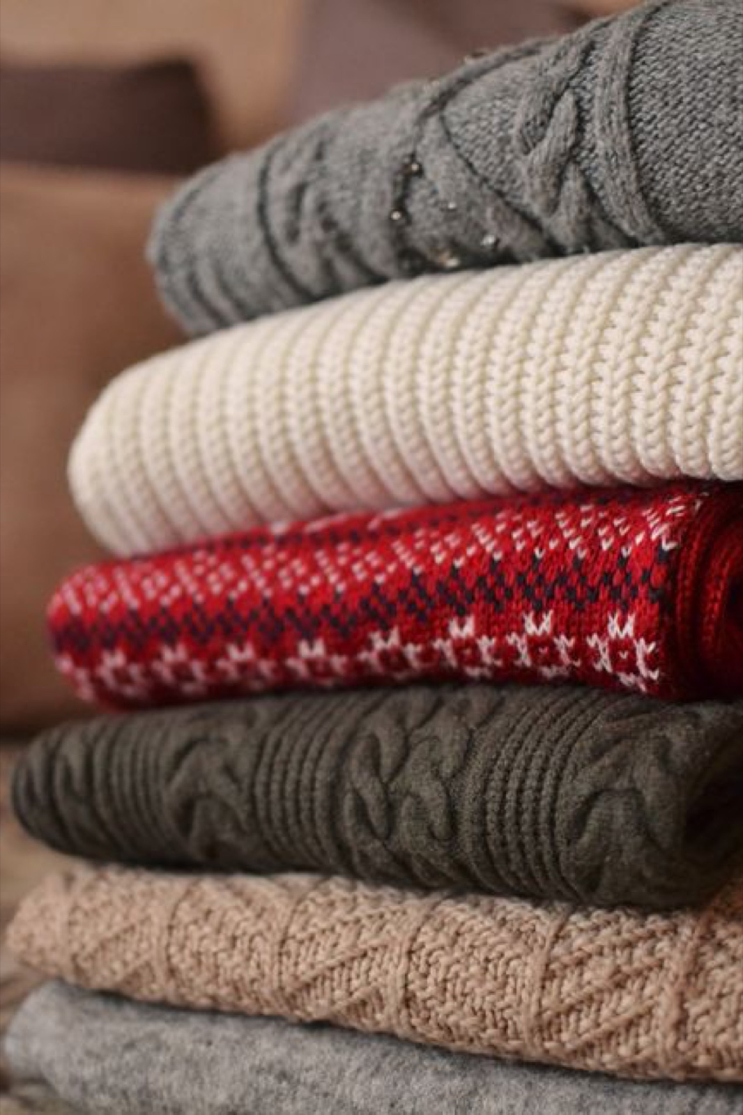 A stacked pile of folded sweaters in gray cable knit, cream ribbed, red fair isle and brown patterned wool