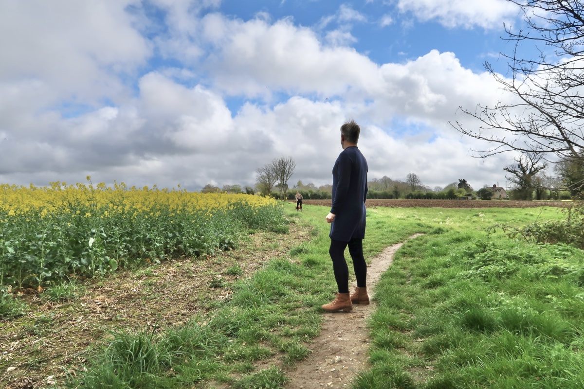 A person seen from behind standing on a grass path beside a yellow rapeseed field, wearing a navy sweater dress, dark tights, and tan suede lace-up boots