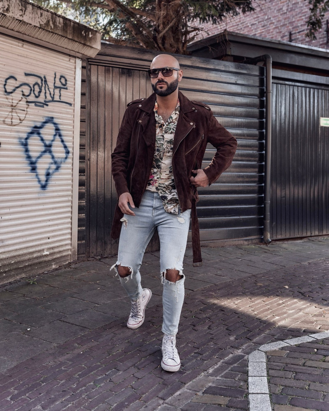 A man in a chocolate brown suede biker jacket and light ripped jeans walking past metal garage doors