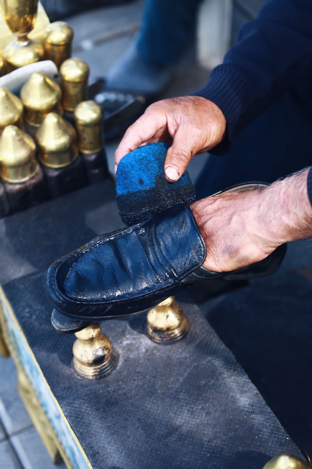 Hands of a shoe shiner applying dark polish to a smooth black leather shoe with a round brush at an outdoor brass-fitted stand