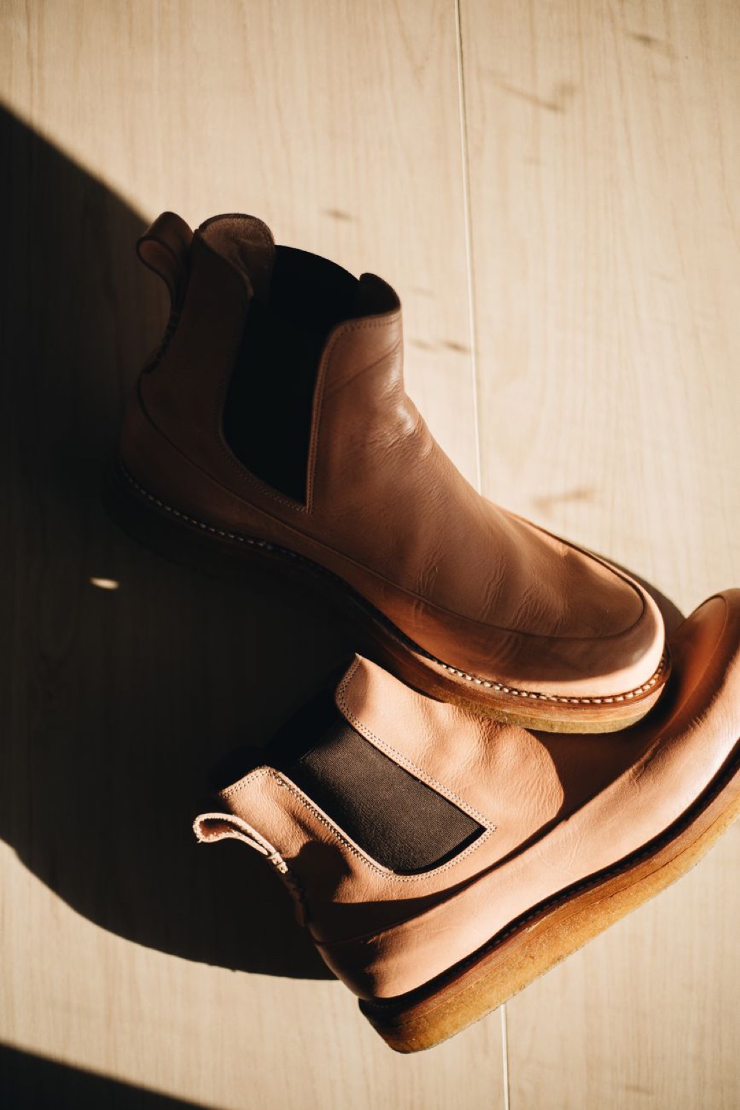 Pair of tan smooth leather chelsea boots with crepe soles lying on a pale wooden floor in hard afternoon sunlight