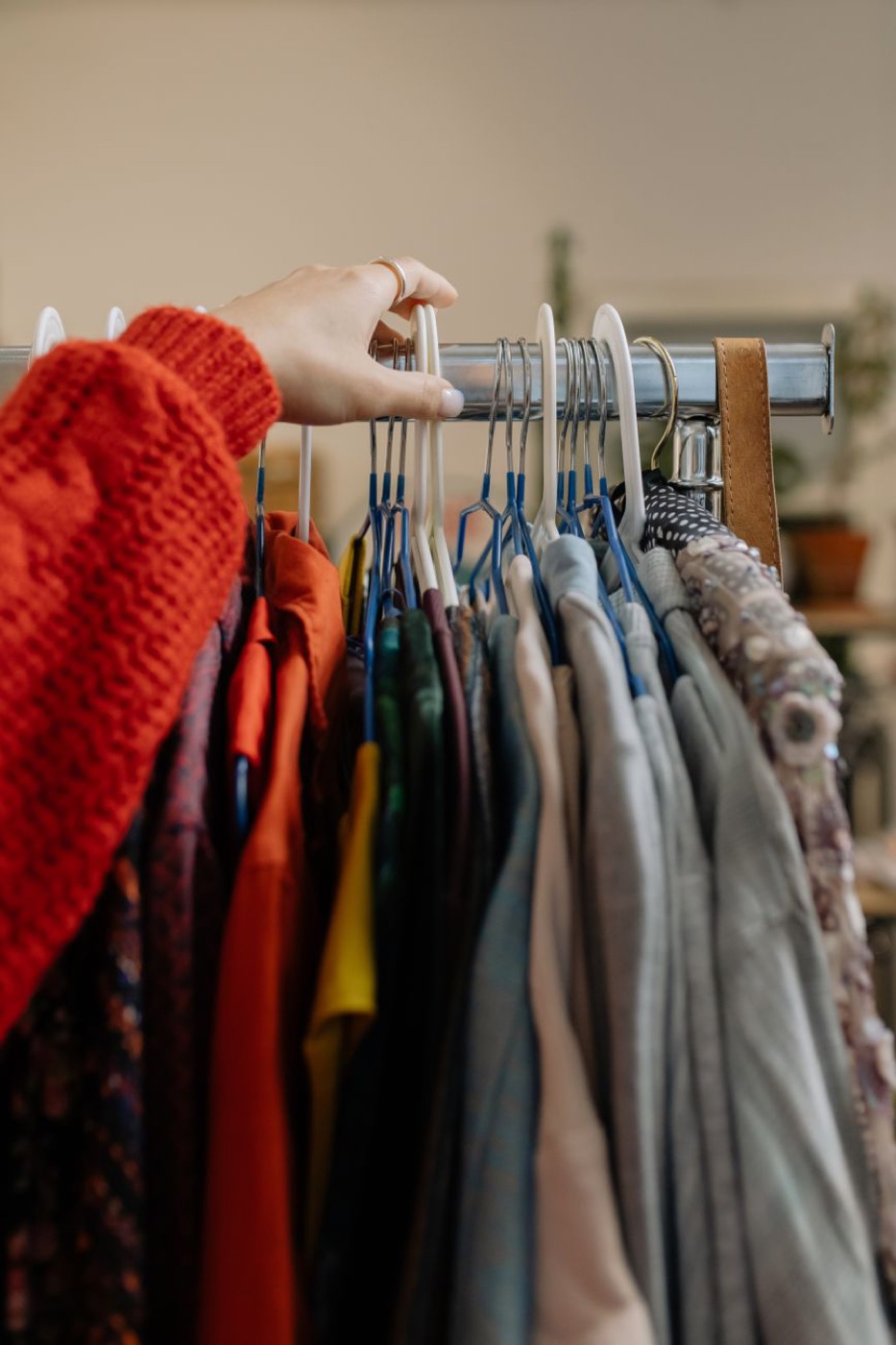 A hand in a red knit sleeve pushing hangers along a chrome rail crowded with shirts and jackets in a small shop