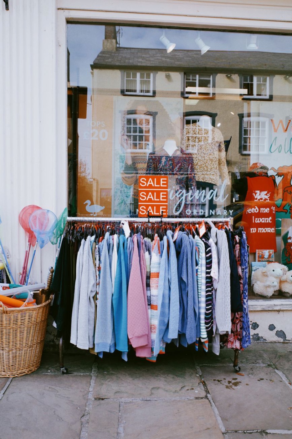 An outdoor rail of knitwear and shirts in pastel colours pushed onto the pavement outside a shop window, with three stacked orange SALE signs behind it