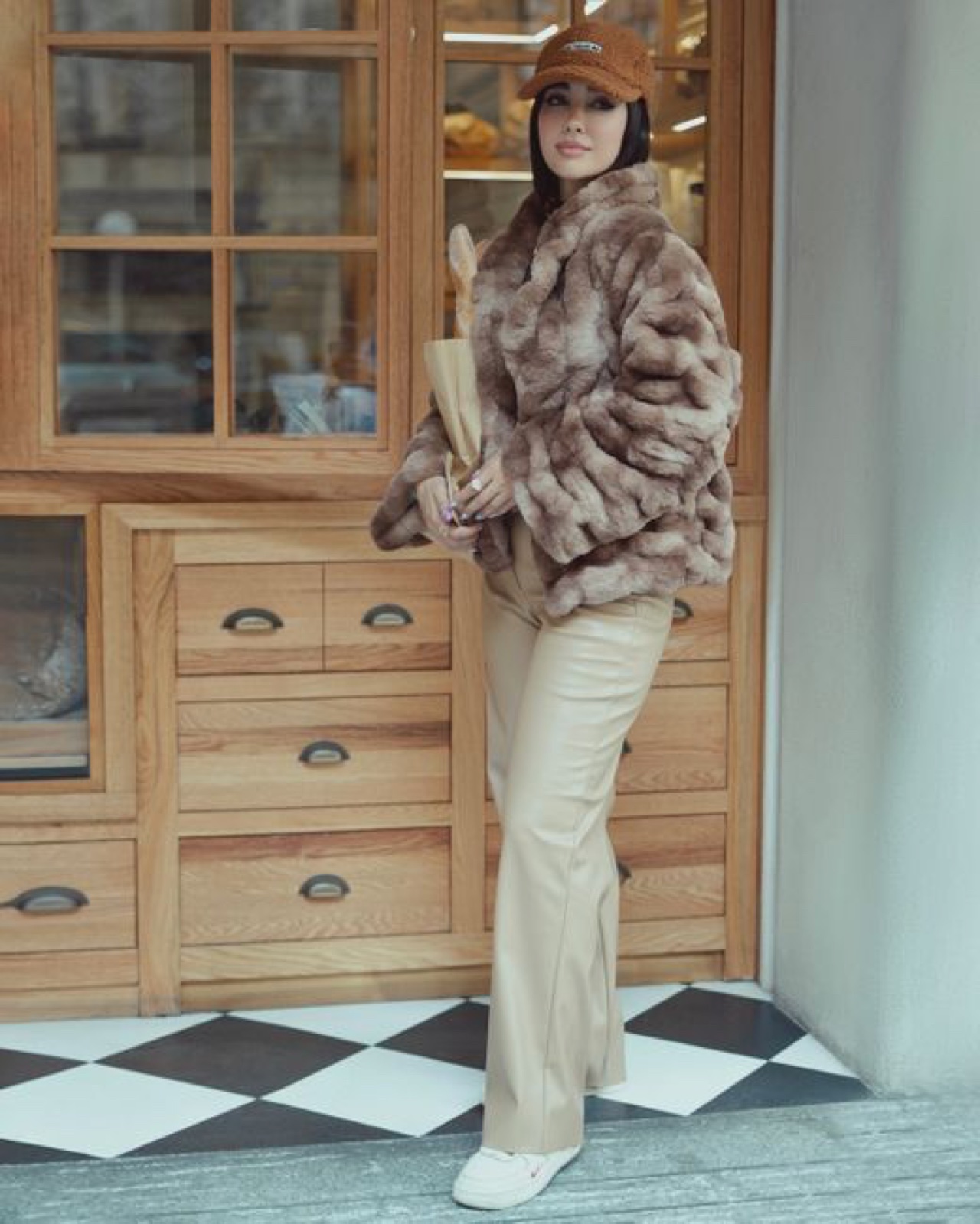 A woman in a pale textured faux fur jacket and brown cap holding a baguette outside a bakery with checkered floor tiles