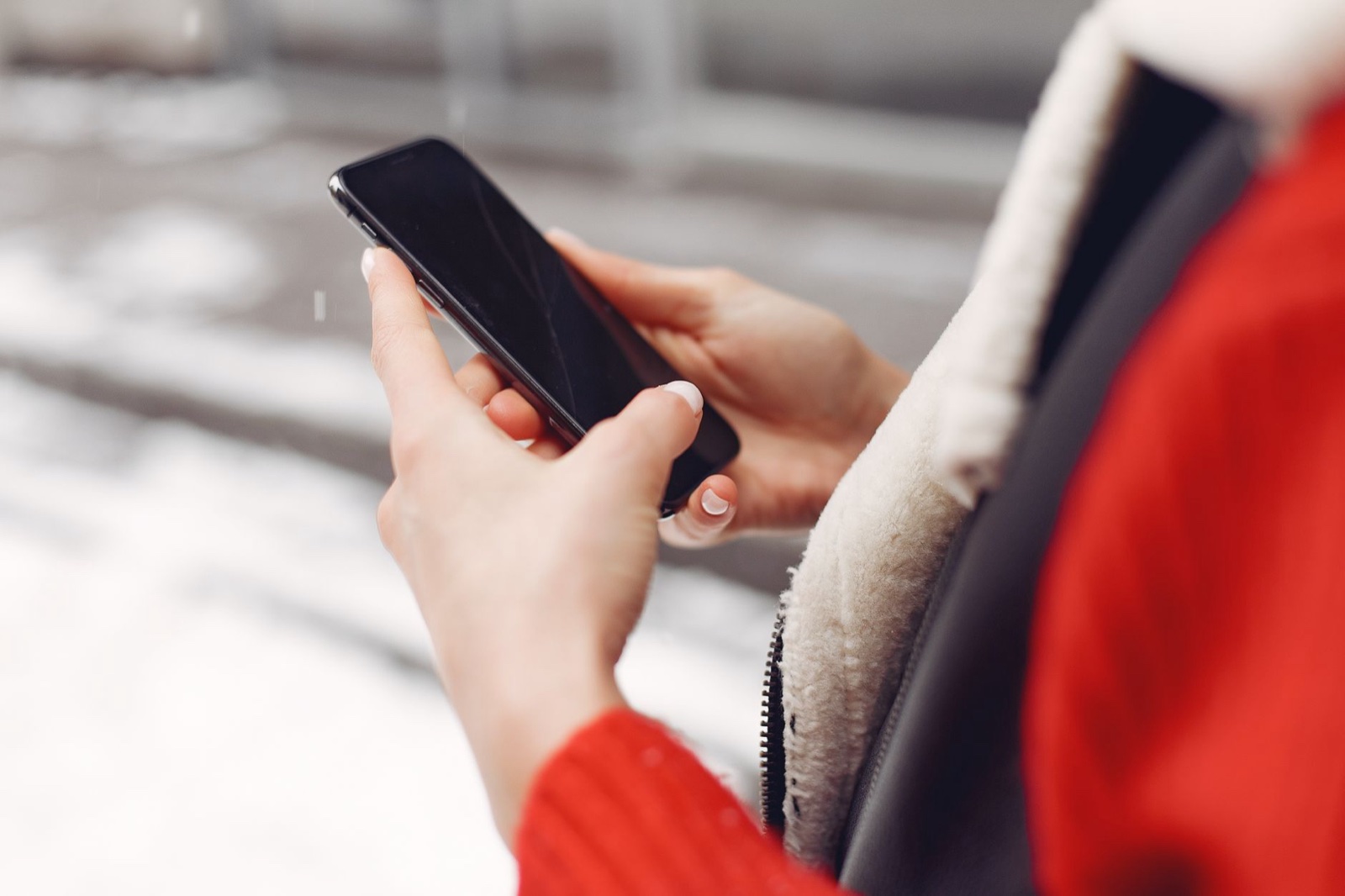 Bare hands holding a smartphone outdoors in winter, wearing a red sweater and shearling-collared jacket, snow visible in the background