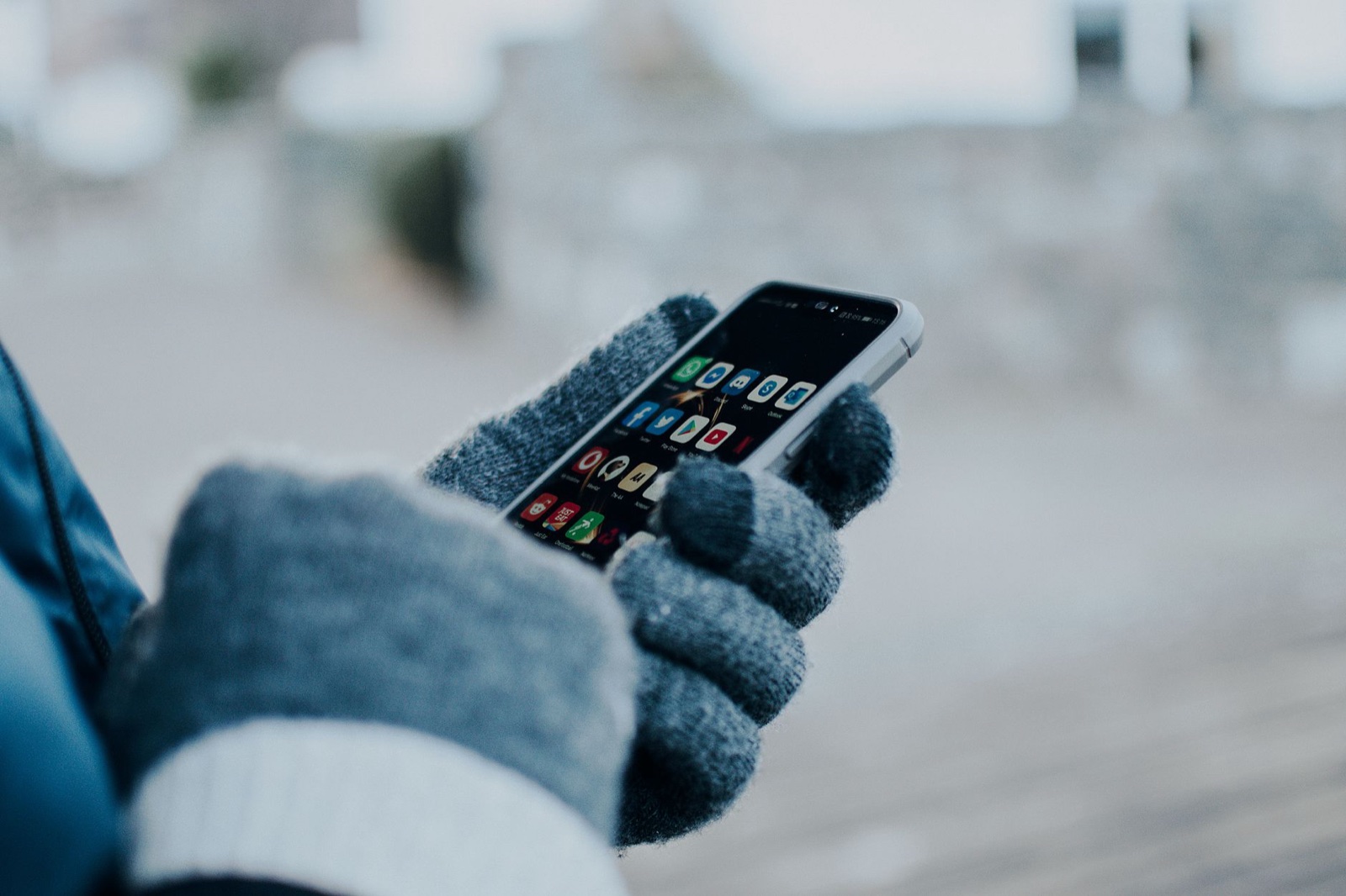 Gray knit gloves with darker conductive fingertip pads holding a smartphone with app icons on screen, photographed outdoors in winter light