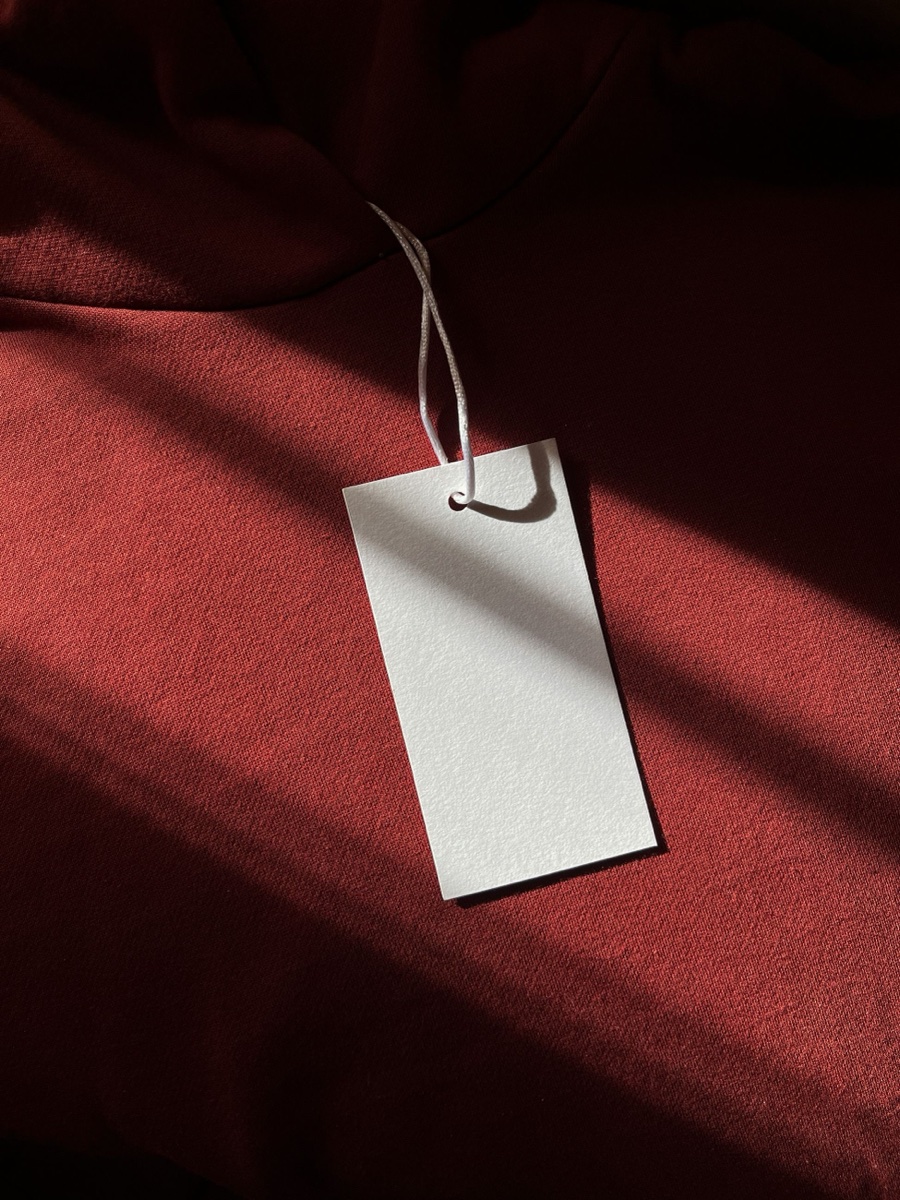 A blank white hangtag on a cord lying across rust-red knit fabric in hard sunlight