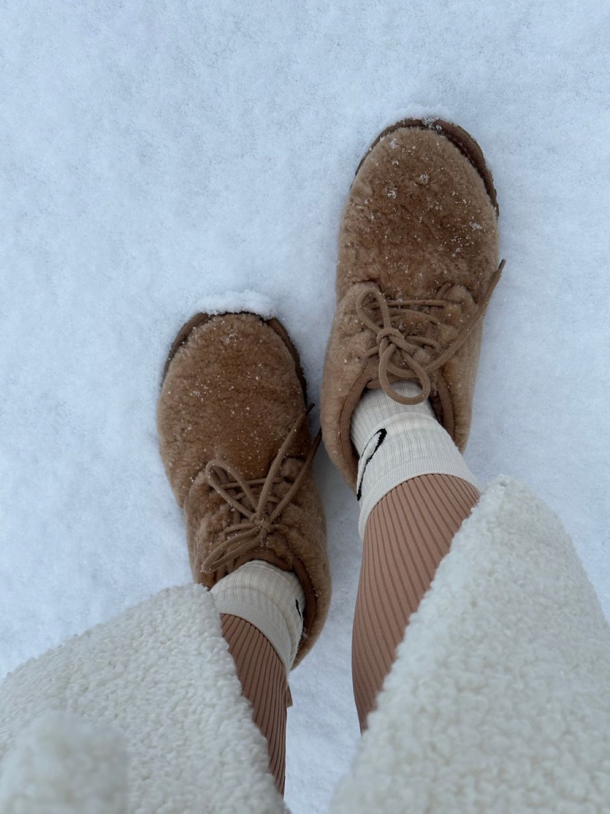 Looking down at a pair of camel-colored curly sheepskin lace-up boots standing in fresh snow, with white fleece pants and ribbed socks