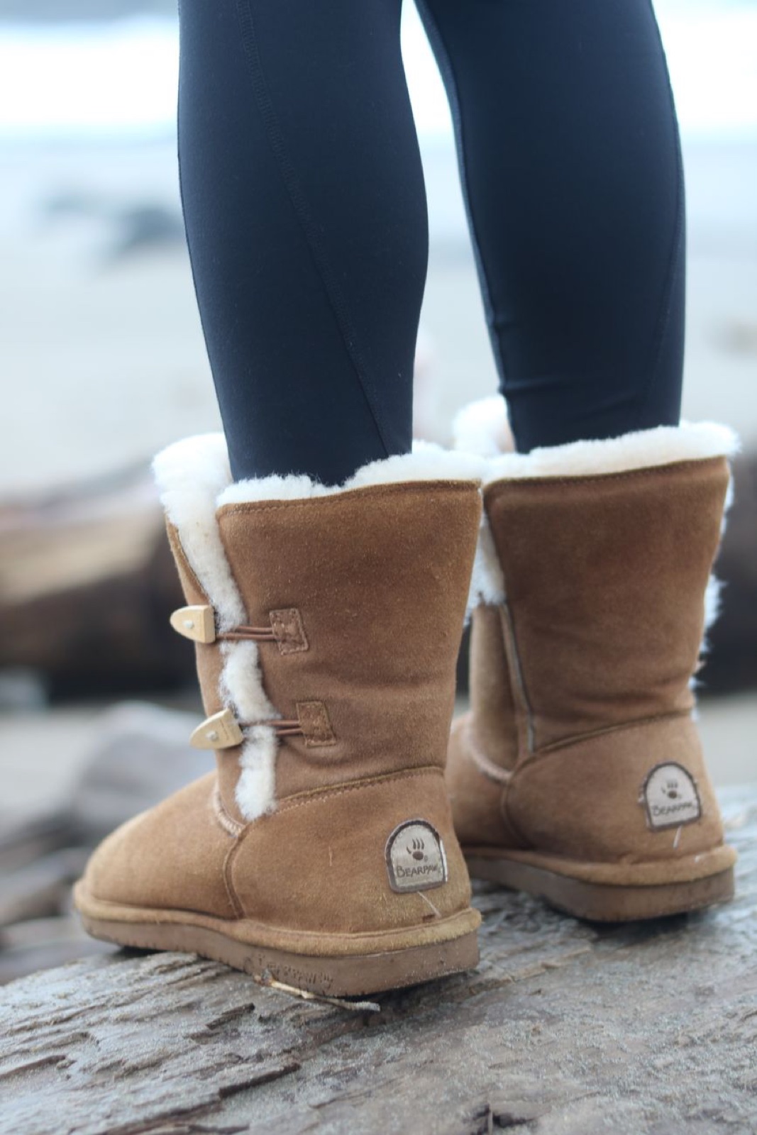 Person in black leggings standing on driftwood at a grey beach wearing chestnut sheepskin-lined toggle boots by Bearpaw