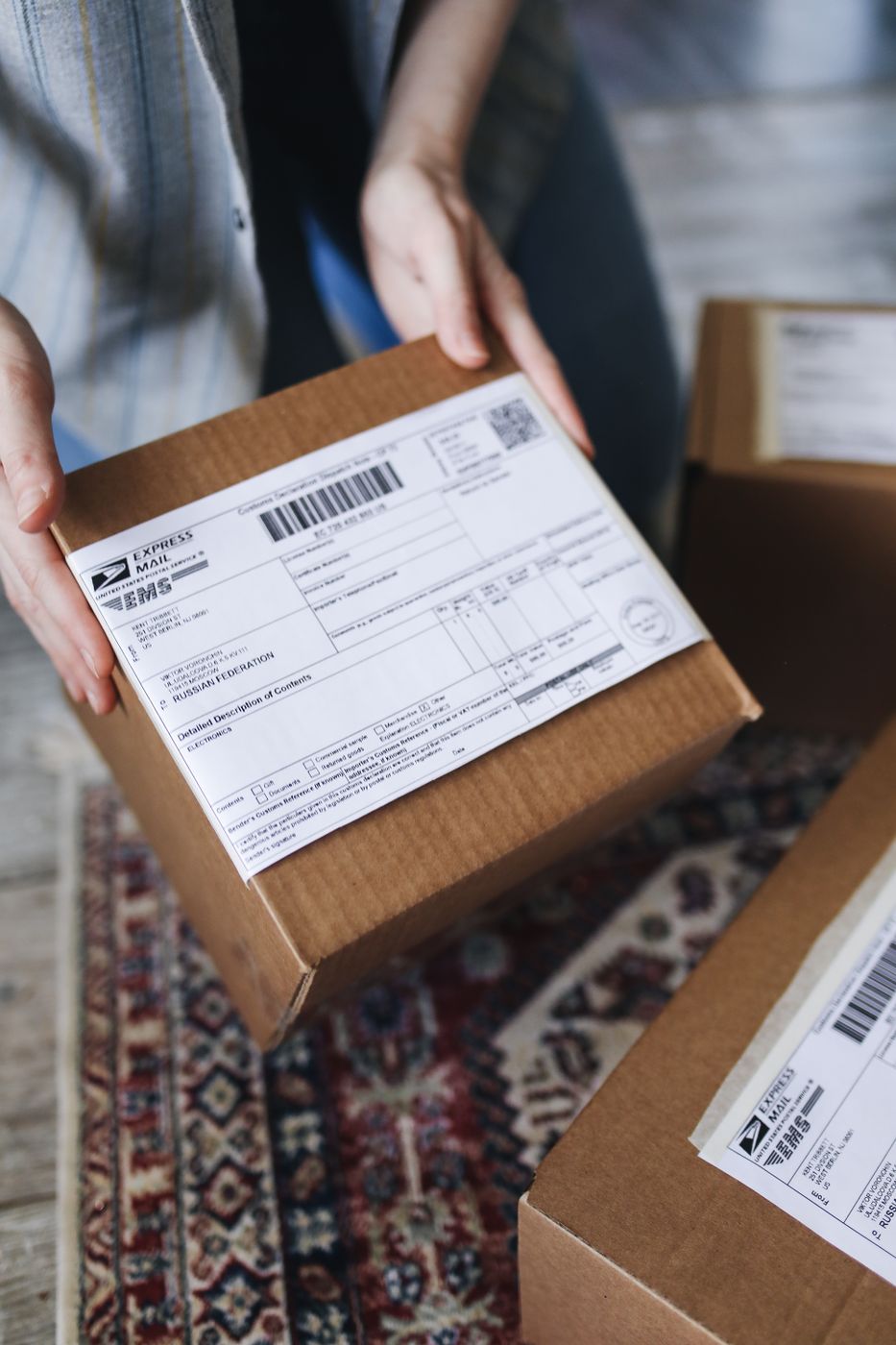 Two hands holding a brown cardboard parcel with a printed shipping label over a patterned rug