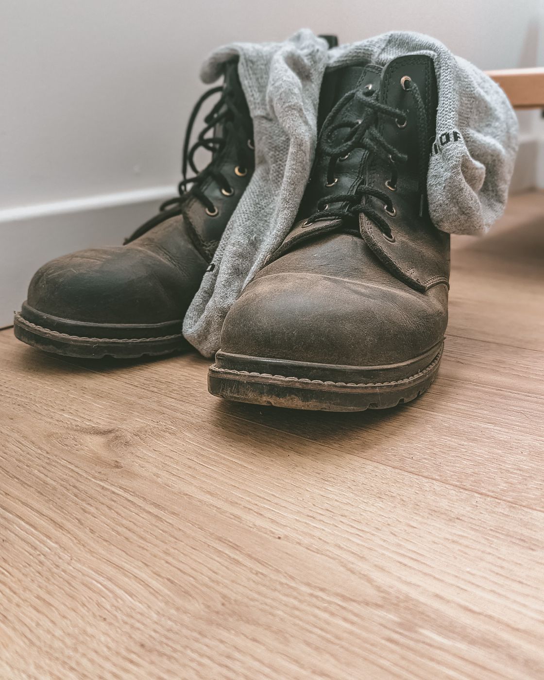 A pair of heavily scuffed dark leather lace-up boots with grey socks tucked in, standing on a pale wood floor