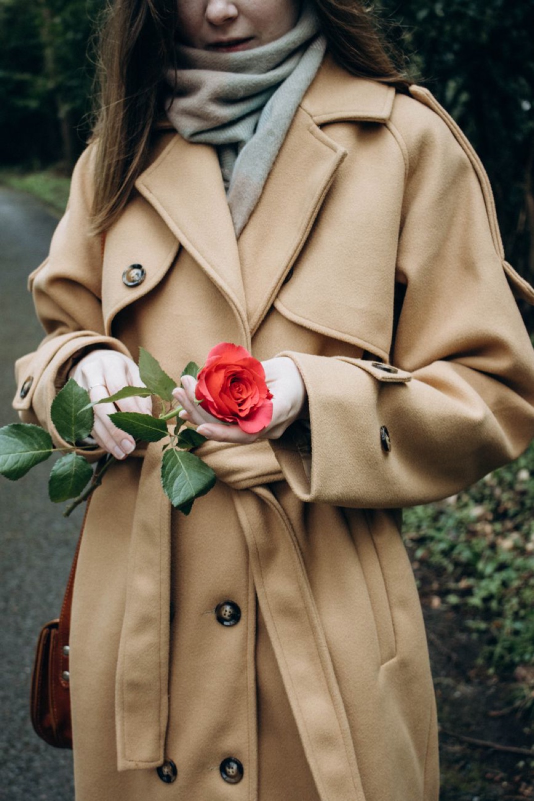 A woman in a pale sandy belted trench coat and sage scarf holding a single red rose, photographed from the chin down on a wet tree-lined path