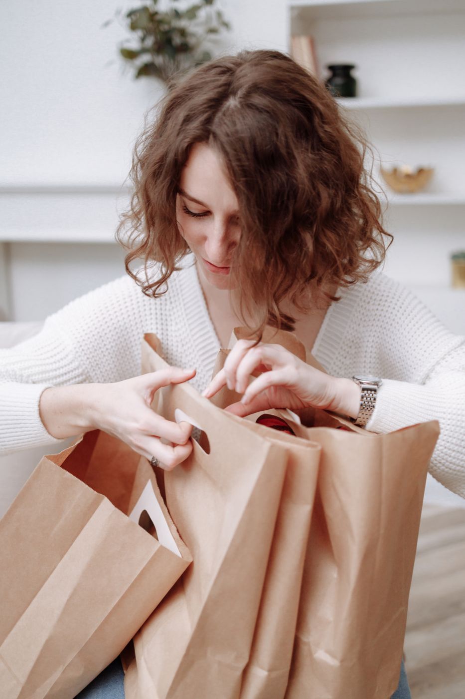 A woman in a white sweater looking down into several brown paper shopping bags indoors